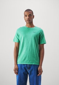 Groene katoenen t-shirt met een ronde hals en korte mouwen. Bevat een klein zwart logo nabij de halslijn, gecombineerd met blauwe spijkerbroeken.
