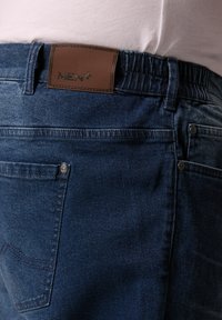 Mörkblå denimjeans med en brun läderlapp märkt "MEN+" vid midjan och en klassisk femfickdesign.