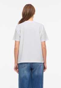 Άτομο με μακριά καστανά μαλλιά που φοράει ένα απλό λευκό κοντομάνικο μπλουζάκι και μπλε τζιν, στέκεται γυρισμένο με την πλάτη σε λευκό φόντο.