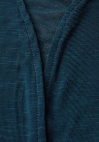 Teal, stickad tyg med en lätt glans. Har en V-ringad design med synlig sömnadsdetalj som skapar en texturerad look.