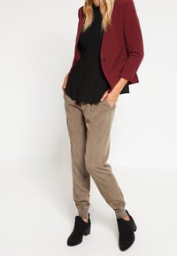 Femme portant un blazer bordeaux, un haut noir, un pantalon jogger taupe et des bottines noires, debout les mains dans les poches sur un fond blanc.