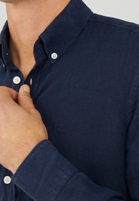 Camicia button-up blu navy con colletto, caratterizzata da bottoni bianchi e un piccolo dettaglio ricamato. Il tessuto presenta un'aspetto texturizzato.