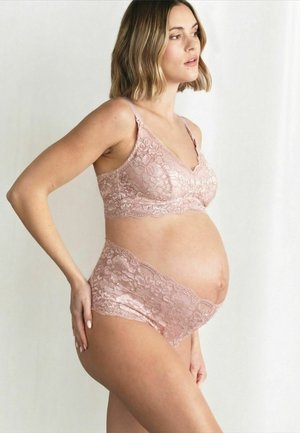 Gravid kvinde med skulderlangt hår iført pink blonde amme-bh og matchende undertøj, stående sideværts med hånd på hoften.