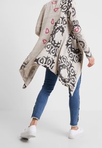 Lichte, beige gebreide asymmetrische cardigan met zwarte en roze bloemmotieven. Gecombineerd met denim jeans en witte schoenen. Lange mouwen.