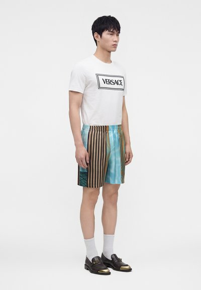 Jeune homme portant un t-shirt blanc Versace, un short à motifs rayés bleu sarcelle et marron, des chaussettes blanches et des mocassins noirs à bouts dorés.