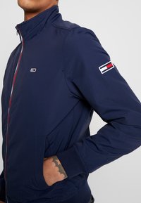 Marinblå bomberjacka i lätt tyg, med hög krage, dragkedja, sidofickor och en logopatch på ärmen.