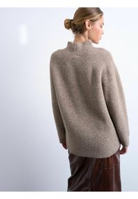 Femme aux cheveux blonds attachés en chignon, portant un pull en maille côtelée beige et une jupe en cuir marron, vue de profil sur un fond clair uni.