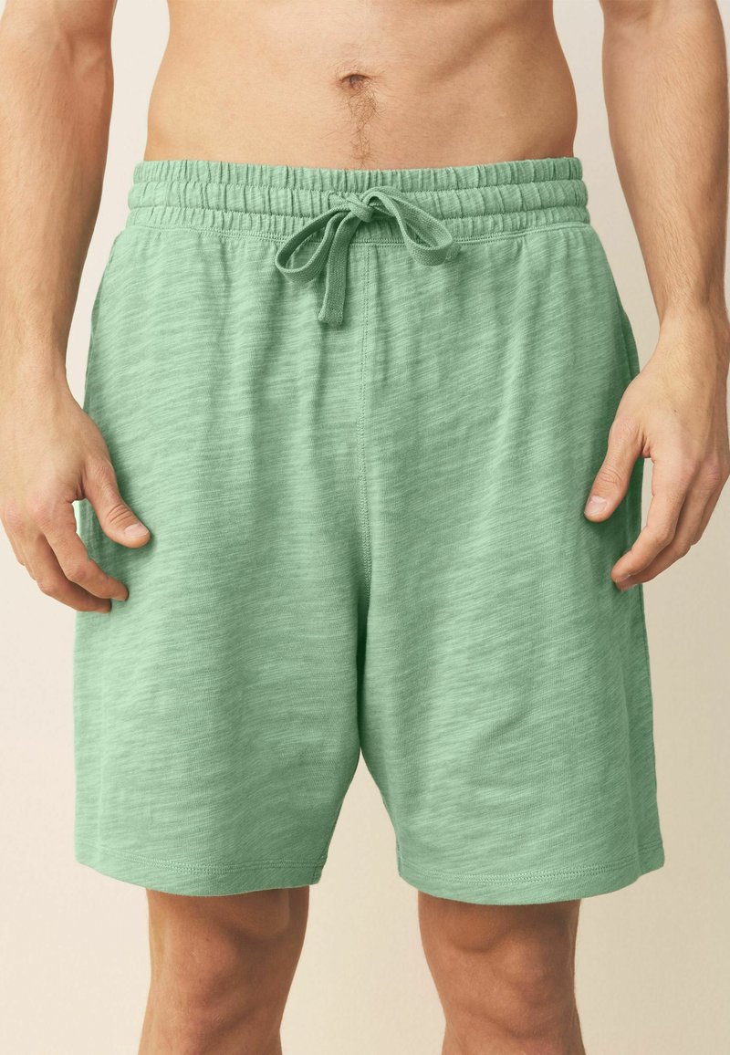 Uomo che indossa pantaloncini verde chiaro con coulisse, in piedi con le mani rilassate lungo i fianchi davanti a uno sfondo neutro.