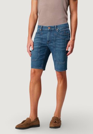 Mężczyzna ubrany w niebieskie szorty dżinsowe z wieloma przednimi kieszeniami, beżowy t-shirt i brązowe mokasyny z frędzlami, stojący na białym tle.