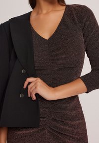 Černý blazer s kulatými klopy, spárovaný s přiléhavými, tmavě lesklými šaty s řasením a výstřihem do V. Měkká textura a elegantní design.