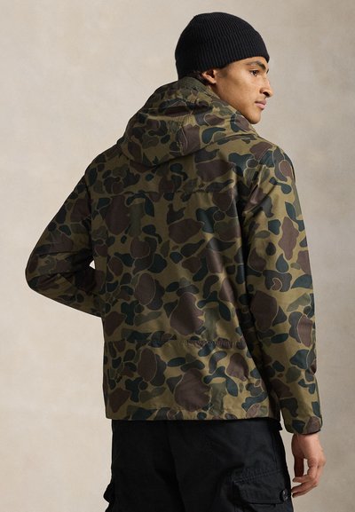 Casaco camuflado em tons de verde e castanho, com capuz e fecho de correr, feito de um material liso e durável. Usado com um gorro preto.