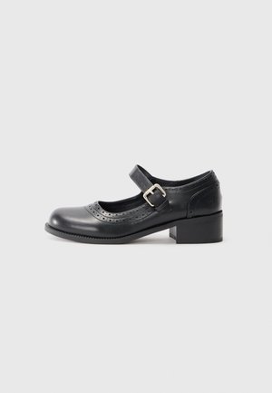 Chaussure Mary Jane en cuir noir avec détail perforé, bride à boucle argentée et petit talon carré sur un fond clair uni.