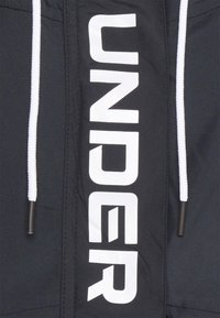 Svart jacka med en framträdande vit "UNDER"-grafik på framsidan, med kontrasterande vita dragsnören och en slät, smidig väv.