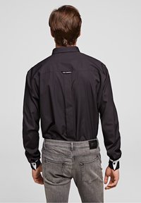 Černá dlouhá rukávová košile s přiléhavým střihem, kontrastními bílými manžetami a štítkem vzadu u límce. Nosená s šedými džíny.