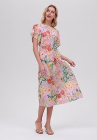 Mekko, jossa on kukkakuosi, pehmeä kangas, lyhyet liehuvat hihat, pastellitausta ja istuva vyötärö midi-pituisena, täydentäen ilmeen nauhattomilla sandaaleilla.