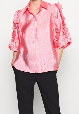 Osoba ubrana w błyszczącą różową koszulę na guziki z falbankami na rękawach i czarne spodnie, stojąca z rękami w kieszeniach na białym tle.