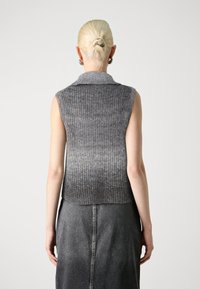 Frau mit blonden Haaren zu einem Dutt frisiert, trägt silberne Creolen, eine ärmellose graue Strickweste und einen ausgebleichten schwarzen Jeansrock, von hinten gesehen.