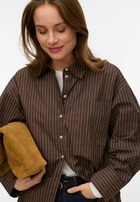 Brązowa koszula na guziki w paski z kołnierzykiem, z białymi paskami, kieszenią na piersi i podwiniętymi rękawami. Towarzyszy jej beżowy materiał.