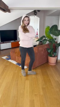 Sweat à capuche rose clair, pantalon slim noir et bottines beiges à la cheville, se tenant dans une pièce avec une plante verte et un canapé brun.