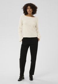 Pull doux de couleur crème avec un col bateau ; associé à un pantalon noir. Le pull a une coupe décontractée et des manches longues.