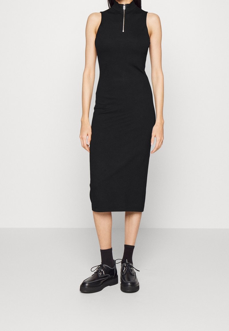 Robe midi noire sans manches avec col zippé, portée avec des chaussettes noires et des chaussures à plateforme lacées, présentée sur une personne debout devant un fond uni.