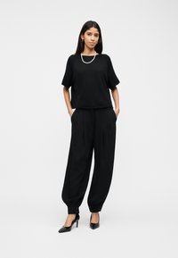 Femme aux longs cheveux foncés portant un pantalon noir ample avec boutons aux poignets, un haut noir à manches courtes, un collier de perles et des talons hauts noirs, debout devant un fond blanc.