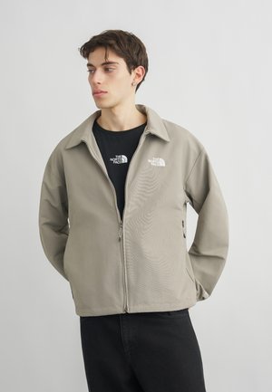 Νεαρός άνδρας που φοράει μπεζ μπουφάν με φερμουάρ και μαύρο t-shirt με λογότυπα "The North Face", στέκεται μπροστά από απλό φωτεινό φόντο.