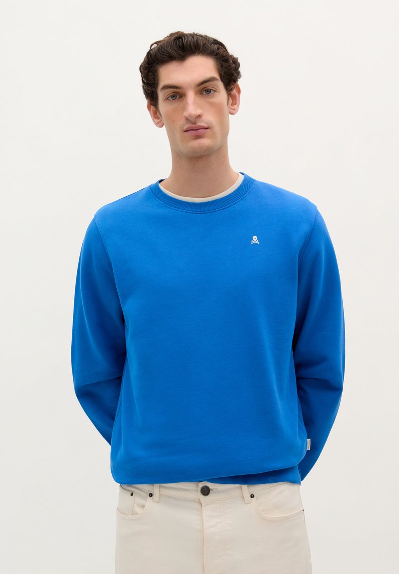 Sweatshirt bleu cu guler rotund, realizat din bumbac, cu mâneci lungi și un logo mic alb pe piept. Purtați cu pantaloni bej.