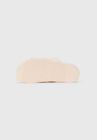 Beige, flauschiger Slide-Sandal mit der Sohle nach oben liegend, die das gerillte Profil zeigt, mit "Steve Madden" in der Mitte eingeprägt.