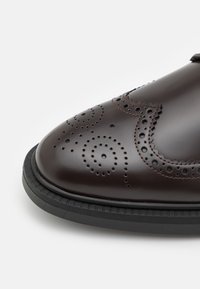 Chaussure en cuir marron avec des motifs tourbillonnants perforés complexes, un bout arrondi et une semelle en caoutchouc noire. Texture lisse avec des coutures contrastées.