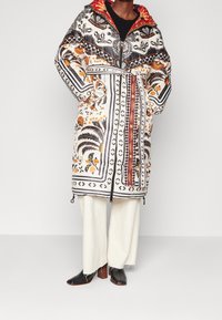 Abrigo largo con capucha de diseño estampado, que presenta motivos florales y geométricos en naranja, negro y crema. Incluye un cinturón y cremalleras en el dobladillo.