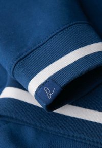 Modrý pletený rukáv svetru s bílým pruhem a malou látkovou nášivkou s vyšitým abstraktním designem smyčky.