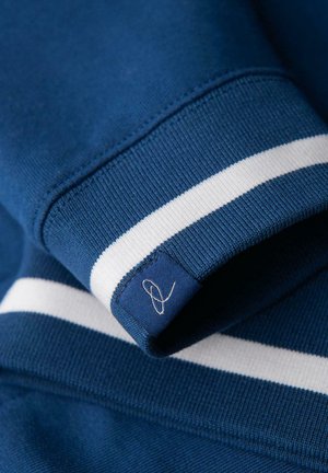 Blauwe gebreide sweatshirtmanchet met witte streep en een klein stoffen label met een geborduurd abstract lusontwerp.