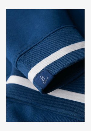 Blauer gestrickter Pulloverbund mit weißem Streifen und kleinem Stoffetikett, das ein gesticktes abstraktes Schleifenmuster zeigt.