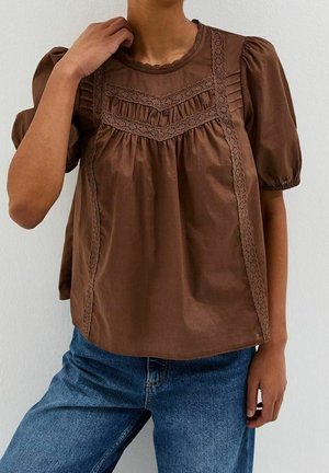 Woman wearing a brown puff-sleeve blouse with lace trim and ruffles paired with blue jeans, posed against a plain wall.