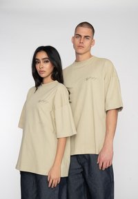 Bežové oversized tričká vyrobené z jemného materiálu, s klesnutými ramenami, okrúhlym výstrihom a jemne vyšitým textom vpredu.