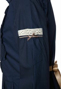 Tengerészkék ing csipkés szegéllyel és barna szalag díszítéssel az ujján, valamint oldalirányú zsebbel, amely hajtásos részletekkel rendelkezik.