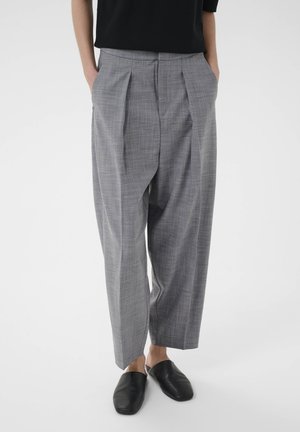 Person wearing gray plaid wide-leg trousers with pleats, hands in pockets, black slip-on shoes, and a black top against a plain background.