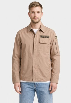 Mężczyzna w beżowej kurtce z kieszenią na piersi i logo „Blauer”, w jasnoniebieskich dżinsach, stoi na tle jednolitego białego tła.