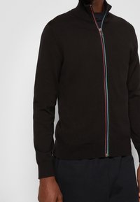 Svart zip-up kofta med hög krage, har en flerfärgad dragkedja. Fitted ärmar och ribbade muddar. Mjuk stickad kvalitet.