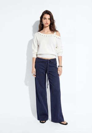 Femme aux longs cheveux bruns portant un pull blanc épaules dénudées et un pantalon large bleu marine, debout devant un fond blanc uni.