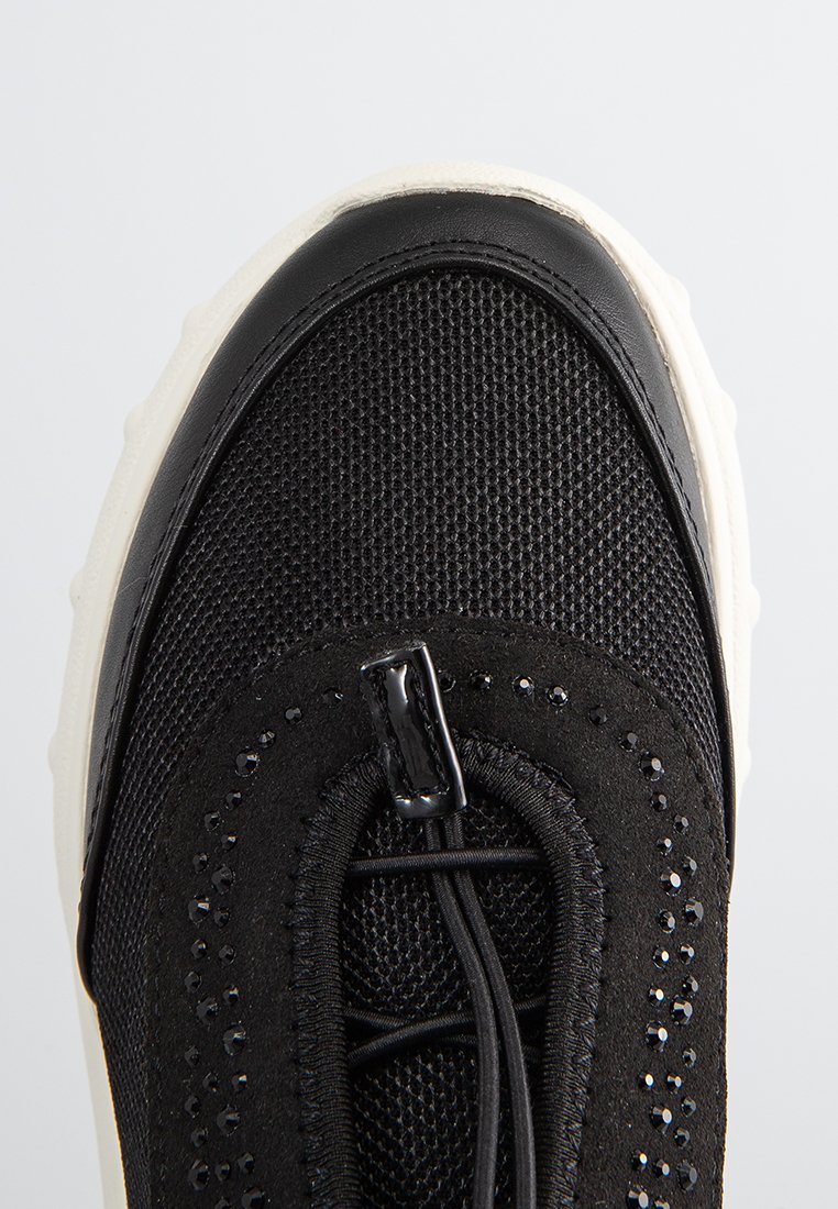 Zapatilla negra con upper de malla, detalles de cuero liso y suela de goma blanca. Incluye acentos decorativos y un sistema de lazada elástica.