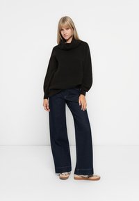 Dunkelbrauner, gestrickter Pullover mit Schalkragen, kombiniert mit weitem, dunklem Jeans. Das Outfit umfasst hellfarbene Schuhe mit einem gemusterten Akzent.