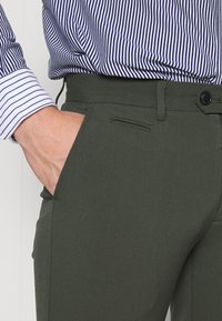 Gröna byxor i släta tyger, med en smal passform, sidoficka och svart knappstängning, tillsammans med en blå och vit randig skjorta.