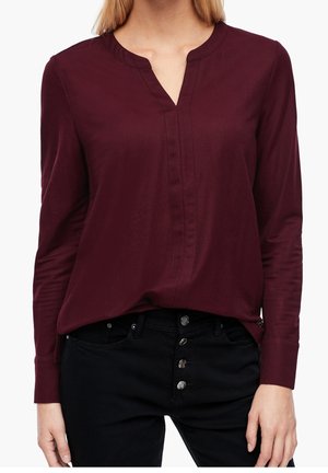 Woman wearing a long-sleeve, dark red blouse with a V-notch neckline and black high-waist buttoned pants.