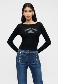 Μαύρο ριμπ μακρυμάνικο μπλουζάκι με στρογγυλή λαιμόκοψη και λευκό τυπωμένο κείμενο. Τζιν παντελόνι ψηλόμεσο με μπλε χρώμα και χρυσά κουμπιά στα πλαϊνά.