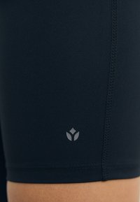 Mørk marineblå treningshorts laget av elastisk stoff med en glatt tekstur. Har en liten, grå logo nederst på kanten og synlige sømmer.