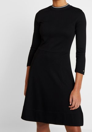 Mujer vestida con un elegante vestido negro ajustado de mangas tres cuartos y un sutil ribete blanco en el escote redondo sobre un fondo liso.