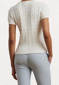 Weißer gestrickter Pullover mit einem Zopfmuster, kurzen Ärmeln und tailliertem Schnitt, kombiniert mit hellgrauen Hosen.