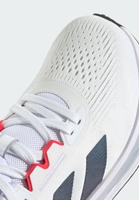 Baskets blanches avec une tige en mesh texturé, des accents rouges et des éléments de design gris, présentant des perforations prononcées pour la respirabilité.